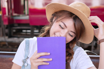 Attractive asian young woman holding book in her hands feeling relaxed, happy. Traveler girl laughing feeling relieve stress during while waiting in the railway station.