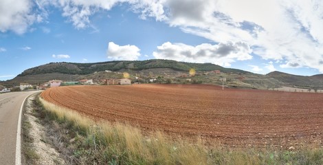 A paved asphalt street next to a plowed field in autumn with some farms on the background among the hills of Monterde rural town in Aragon, Spain