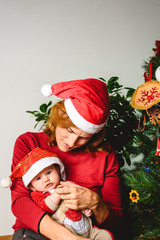 Baby girl dressed as Santa Claus lovingly embraced by her protective mother, family concept at Christmas.