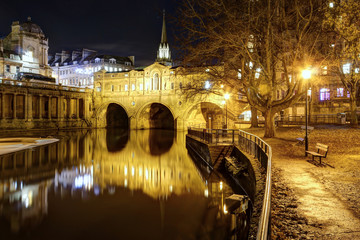 The Pultney bridge reflecting into the Avon river next to a park in winter, with night lights, in Bath, Somerset region, United Kingdom