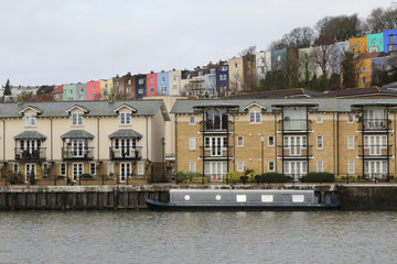 A moored flatboat in the Avon river wharf, in front of beige and colored terraced houses, during winter, in Bristol, United Kingdom