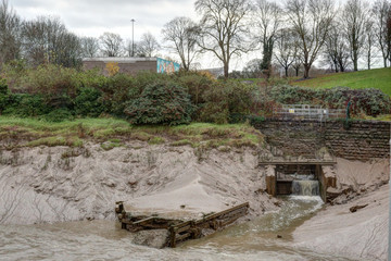 A creek flowing into the Avon river, surrounded by beige and grey mud and a green lawn, with bare trees in a cloudy winter in Bristol, United Kingdom