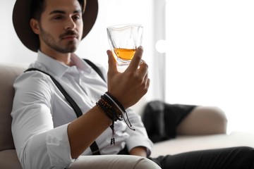 Young man with glass of whiskey sitting on sofa at home. Space for text