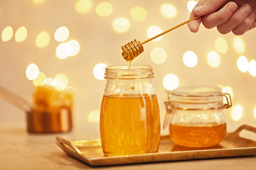 Woman holding dipper over jar with honey on table against blurred lights