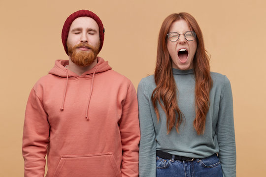 Indoor shot of young ginger beard male keeps his eye close and keep calm. Young redhead woman open mouth wide and screams, feels andgry and mad about some problems