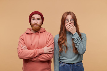 indoor shot of young ginger couple, dressed casualy, looks into camera with shocked facial expression, great surprisement, close mouth with hand. Isolated over biege background
