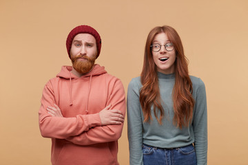 Indoor shot of young ginger female with long hair, wears casualy, smiles and looks aside to her friend standing near her. Man keeps hand folded, raise eyebrow and have astonished facial expression.
