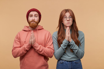 Young attractive ginger couple posing against biege background, dressed casualy and keep hands in praying gesture.