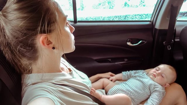 Woman is sitting in the car in the back seat of a car with a sleeping baby in her laps