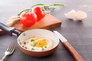 Fried egg in a pan, tomatoes and green onions are cooked for breakfast on a wooden gray table.