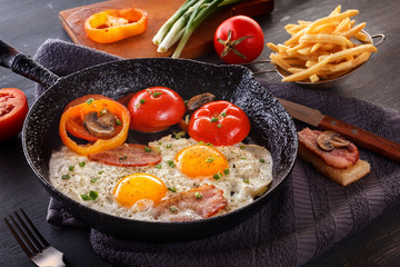 Fried eggs with bacon and tomatoes on an old cast-iron pan with french fries on a gray table. Close-up
