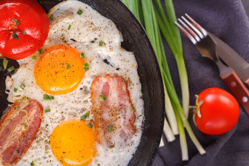 Fried eggs with bacon and tomatoes on an old cast iron pan and cutlery on a gray table. Close-up. Top view