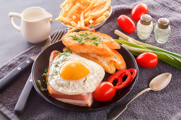 Fried egg with bacon in a black plate with fried pieces of bread, greens tomatoes, a jug of milk and French fries on a gray wooden table. Close-up