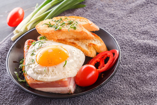Fried egg with bacon in a black plate with fried pieces of bread, greens and tomatoes on a gray wooden table. Close-up. Copy space