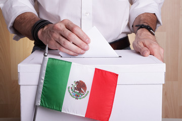 A Mexican citizen inserting a ballot into a ballot box. Mexico flag in front of it