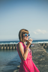 Young woman in a long red dress and with a glass of wine posing on the sea background.