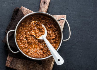 Homemade beef bolognese sauce in a saucepan on a wooden chopping board, on a dark background. Traditional Italian ingredient for pasta