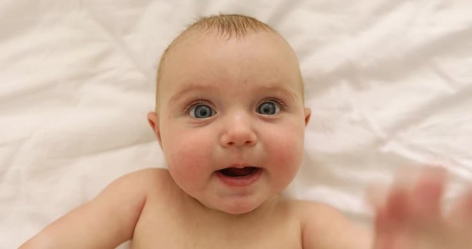 From above shot of charming little baby on white bed looking at camera and showing tongue