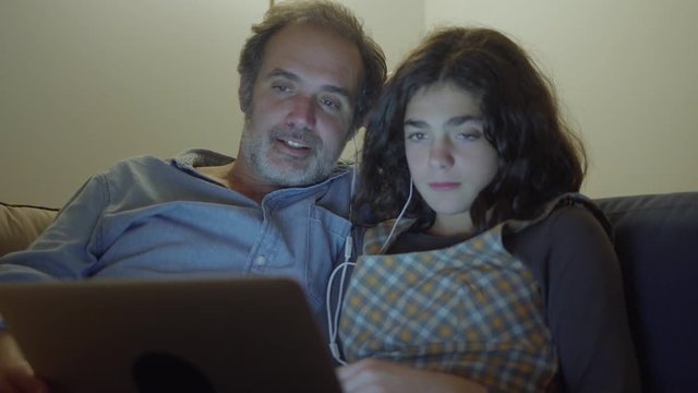 Concentrated father and daughter sitting on couch and watching movie. They watching feature on laptop, they both wearing earphone to keep volume down. Free time concept.