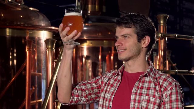 Cheerful male brewer smiling to the camera, examining beer in a glass. Brewery worker standing in front of metal containers at brewery, holding glass of beer for examination