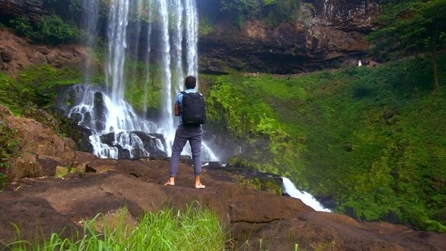 backside guy with backpack stands on rock watches waterfall