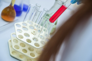 A man's hand hold and lean a glass test tube(Proberohr) with red(magenta) water(liquid, fluid) on the reflected desk in the laboratory