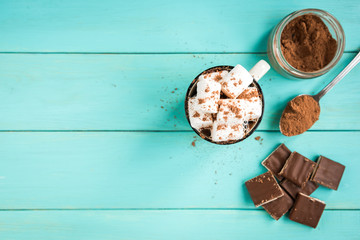 A cup of coffee with marshmallow, a knitted blue scarf, chocolate bar and cocoa on green wooden background. Drinks and sweets.