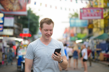 Young happy tourist man smiling while using phone against view of Khao San road in Bangkok