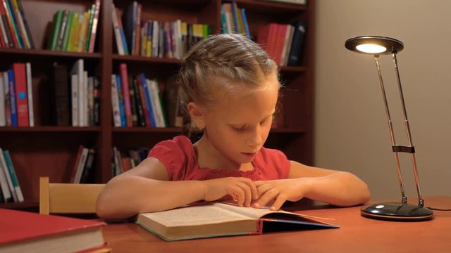 six year old girl reading sitting at the table by the light of a desk lamp