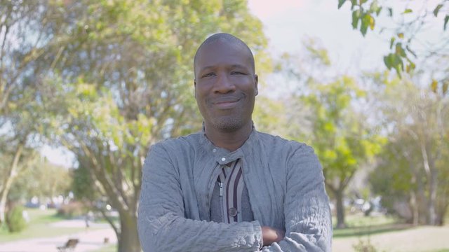 Smiling African American man pising with crossed arms in park. Head and shoulders portrait of handsome man in summer park. Joy concept