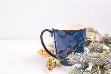 Dark blue mug with hot tea, coffee or cocoa, decorative golden cones and fir tree branches on white textured  background.