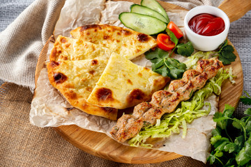 Traditional georgian food - shish kebab and khachapuri pie served with fresh vegetables at wooden tray board on table with sack cloth background.