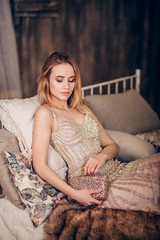 Fashion photo of a luxury beautiful blonde woman in a long stylish Golden dress and a clutch on her hand on the couch during the party. Celebrating birthday or new year