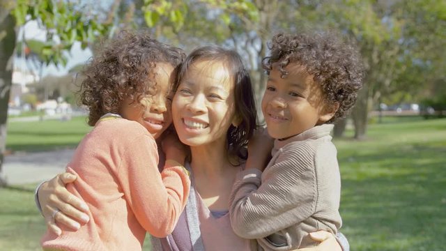 Happy African American kids kissing and hugging Asian mother. Smiling multicultural family embracing in summer park. Family concept