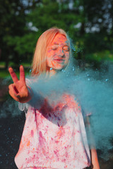 Magnificent blonde model playing with exploding blue dry powder and showing peace gesture