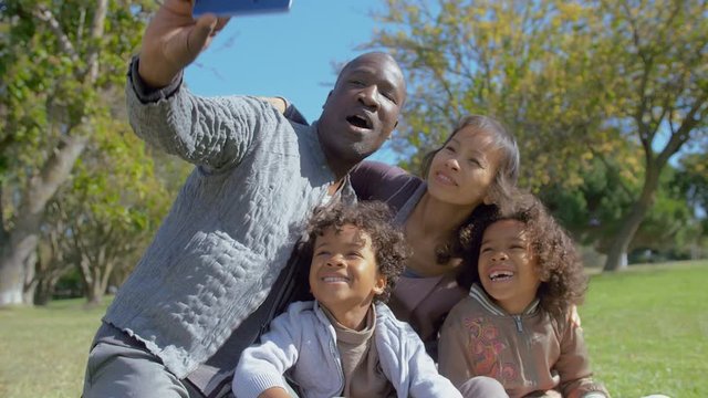 Happy Multiethnic family taking selfie on smartphone in summer park in slow motion. Interracial family with kids having video call on digital device. Communication concept