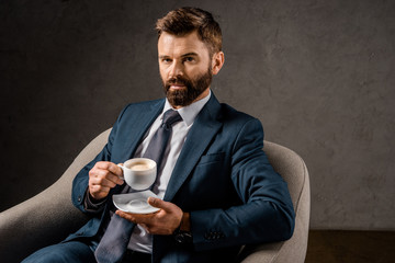 serious businessman showing white cup with coffee while sitting in armchair