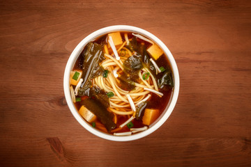 An overhead photo of a bowl of miso soup with tofu, scallions, noodles, wakame seaweed and enoki mushrooms on a dark wooden background with copy space