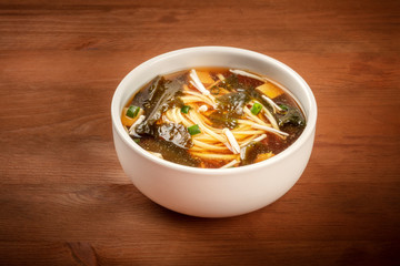 A photo of a bowl of miso soup with tofu, scallions, noodles, wakame seaweed and enoki mushrooms on a dark wooden background with a place for text