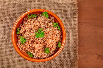 Cooked buckwheat in an earthenware bowl, garnished with fresh parsley leaves, shot from the top on a burlap on a dark rustic wooden background with a place for text