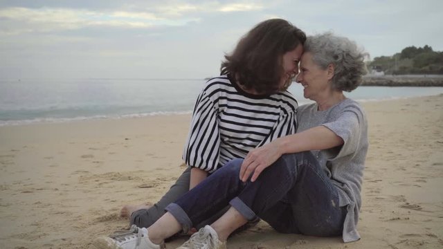 Senior mother and happy daughter touching noses on beach and having fun. Cheerful women laughing while sitting on sand beach near the sea. Togetherness concept