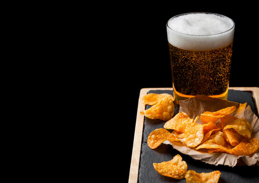 Glass of lager beer with potato crisps snack on stone board on black background. Beer and snack. Space for text