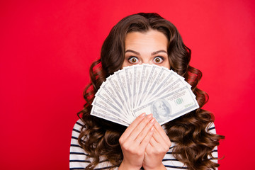 Portrait of nice funny attractive frightened wavy-haired lady wearing striped pullover holding in hands hiding behind fan of hundreds usd dollars isolated over bright vivid shine red background