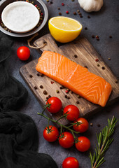 Fresh raw salmon slice on chopping board with pepper tomatoes and lemon on stone kitchen table background. With rosemary and salt
