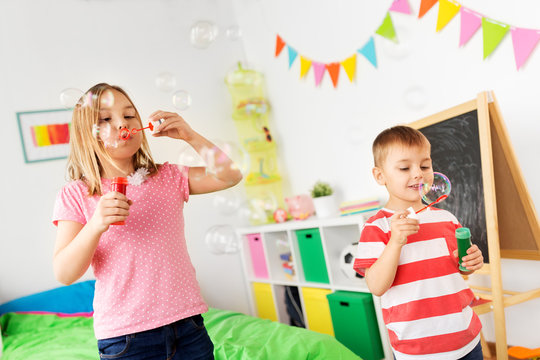 childhood, leisure and people concept - happy children blowing soap bubbles at home