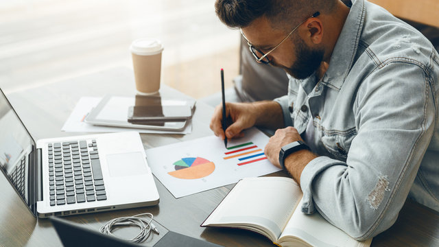 Bearded hipster man sits at table,working on laptop, and makes notes in chart, graph, diagram.Businessman analyzes data.