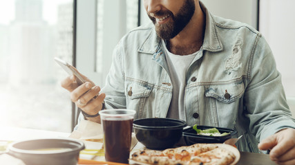 Hipster guy sits in cafe at table, has lunch, using smartphone. Man pays order in restaurant using...