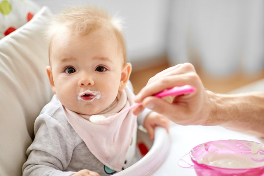 family, food, eating and people concept - father feeding little baby sitting in highchair with puree by spoon at home