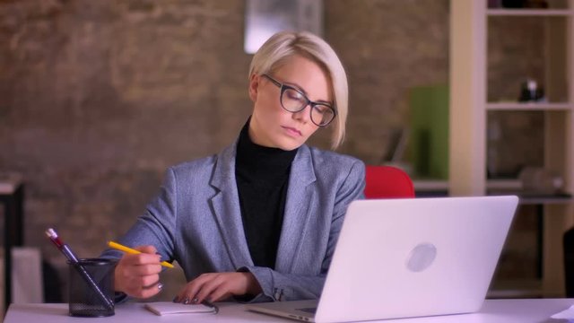Portrait of blonde short-haired businesswoman in glasses working with laptop and making notes into the notebook in office.