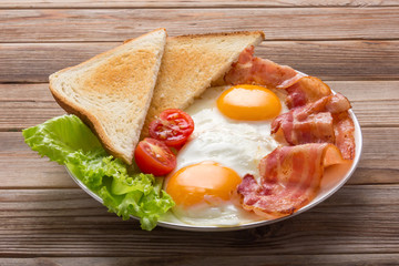 white plate of fried eggs with bacon, toasts and vegetables on wooden table, top view